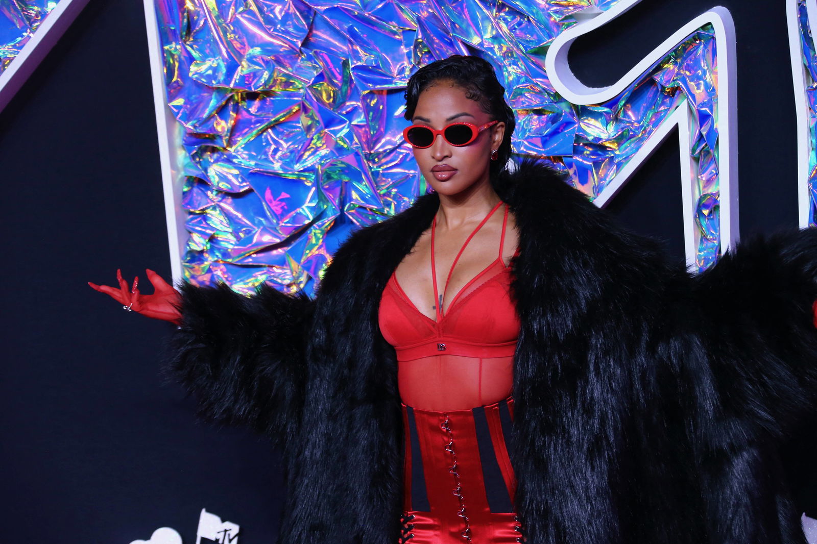 Dancehall, Rap und Reggae Musikerin Shenseea vor einer Wand in einem Roten Korsett, darüber ein schwarzer Pelzmantel. Zudem trägt sie eine Brille mit rotem Rand.