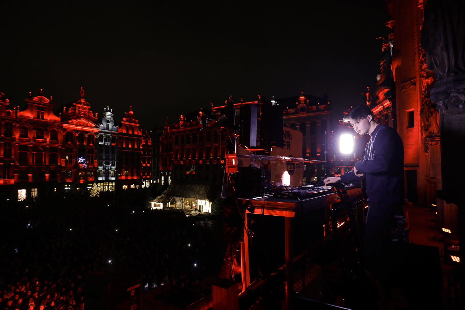 DJ Petit Biscuit in einem lilanen Pullover am Mischpult auf einer Balkonartigenerhöhung. Im Hintergrund rot angeleuchtete Altbaugebäude.